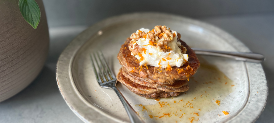 Stack of carrot cake pancakes topped with cream cheese frosting, walnuts, orange zest and maple syrup on a plate